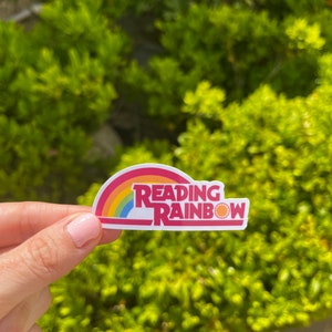 May include: A white sticker with a rainbow arc and the text "Reading Rainbow" in red and yellow letters. The rainbow has red, orange, yellow, green, blue, indigo, and violet stripes. A small yellow sun is at the end of the rainbow.