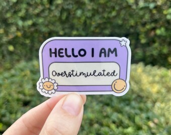 May include: Purple and white sticker with the text "Hello I am Overstimulated" in black cursive font. The sticker has a white star and two smiley faces on either side of the text.