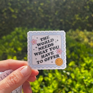 May include: A blue and white sticker with a white border and a white star pattern. The sticker has the text "The world needs what you have to offer" in black text with a pink flower and a yellow smiley face.
