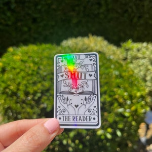 May include: A holographic sticker with a black and white illustration of a book, hands, and the words "SMUT THE READER" with the number "XVIII" above the book. The sticker has a rainbow iridescent background.