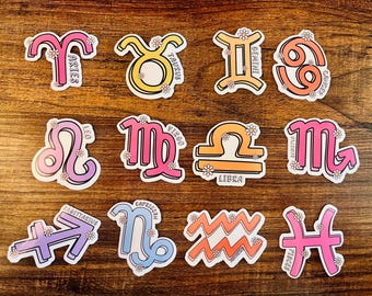 May include: Twelve colorful zodiac sign stickers with white flower accents. Each sticker features a different zodiac symbol and its corresponding name. The stickers are on a wooden surface.