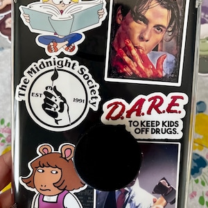 May include: A black phone case with various colorful stickers. The stickers include a cartoon character reading a book, a Scream movie poster, a logo for "The Midnight Society" with the text "EST 1991", a red sticker with the text "DARE TO KEEP KIDS OFF DRUGS", and a cartoon character with brown hair and a pink shirt.