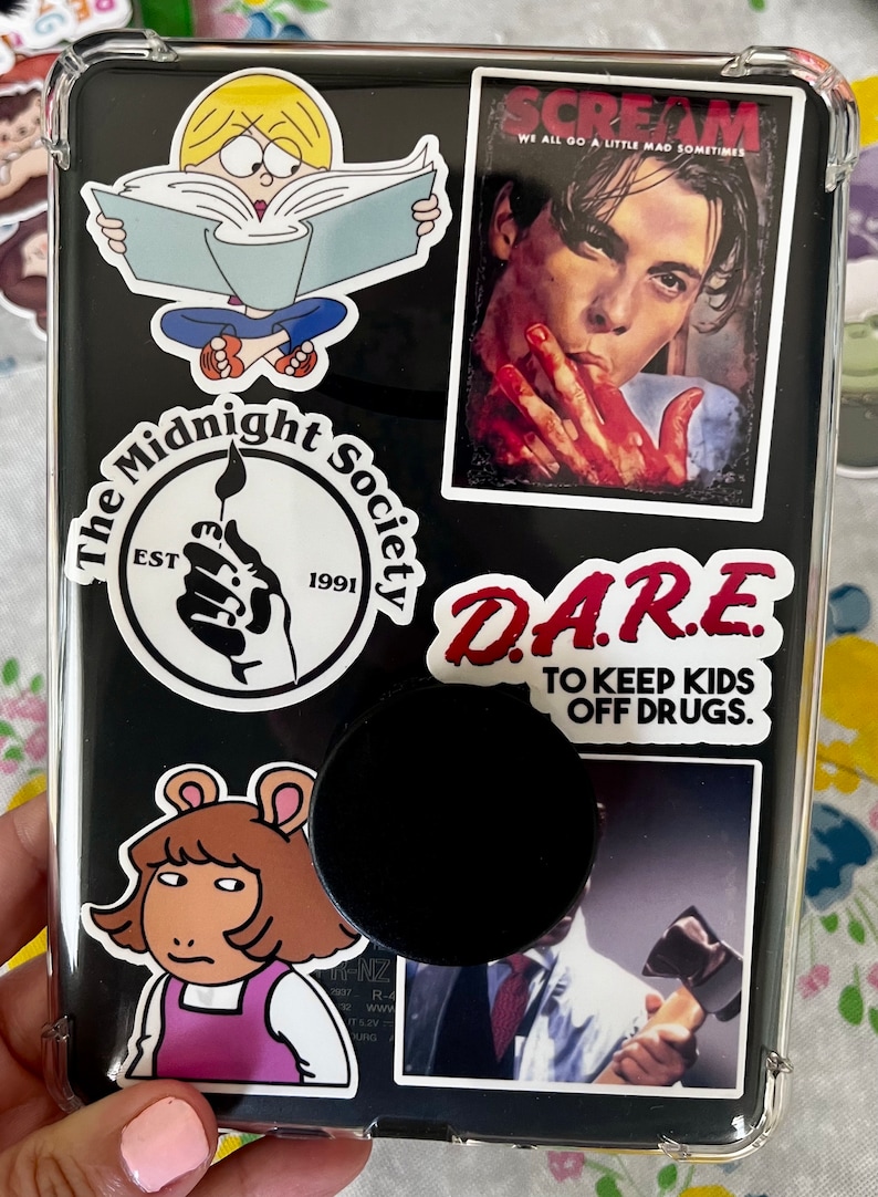 May include: A black phone case with various colorful stickers. The stickers include a cartoon character reading a book, a Scream movie poster, a logo for "The Midnight Society" with the text "EST 1991", a red sticker with the text "DARE TO KEEP KIDS OFF DRUGS", and a cartoon character with brown hair and a pink shirt.