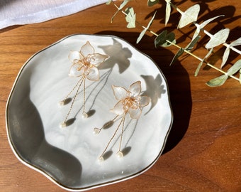 May include: A pair of delicate white flower earrings with gold wire and pearl accents. The earrings are displayed on a white plate with a wooden background.