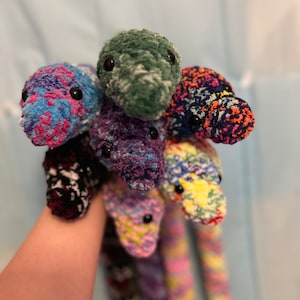 May include: A collection of colorful crocheted snake toys. The snakes are made with different colored yarns and have black eyes.