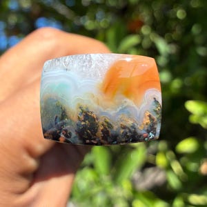 May include: A polished rectangular agate gemstone featuring a landscape-like pattern with a translucent white top, a vibrant orange section, and a base filled with dark, mossy green and black dendritic inclusions layered in wavy bands.