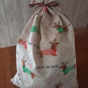 May include: A beige fabric gift bag with a brown drawstring closure. The bag is patterned with cartoon dachshunds wearing sweaters and reindeer antlers, candy canes, and the words "Happy Holidays".