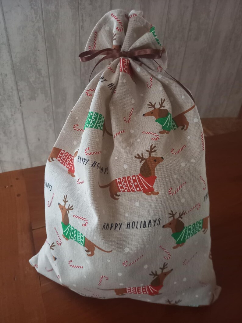 May include: A beige fabric gift bag with a brown drawstring closure. The bag is patterned with cartoon dachshunds wearing sweaters and reindeer antlers, candy canes, and the words "Happy Holidays".