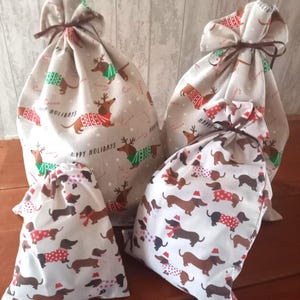 May include: A collection of four drawstring gift bags with a festive dachshund pattern. The bags feature dachshunds wearing holiday sweaters and hats, with the words "Happy Holidays" printed on the fabric. The bags are tied with brown ribbons.
