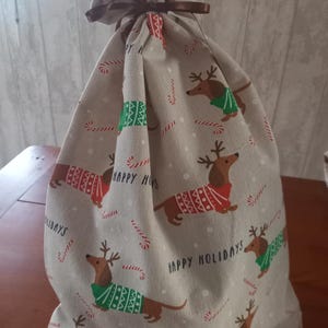 May include: A beige fabric gift bag with a brown ribbon closure. The bag is patterned with dachshunds wearing holiday sweaters and reindeer antlers, candy canes, and the words "Happy Holidays".