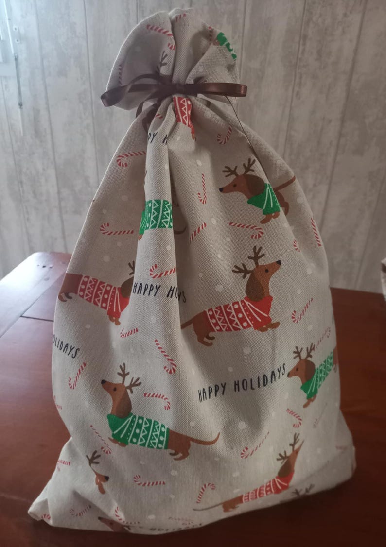 May include: A beige fabric gift bag with a brown ribbon closure. The bag is patterned with dachshunds wearing holiday sweaters and reindeer antlers, candy canes, and the words "Happy Holidays".