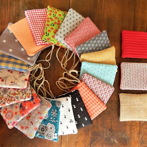 Peut inclure: Une collection de 16 pochettes en tissu colorées avec différents motifs, disposées en cercle sur une surface en bois. Chaque pochette a une fermeture à cordon et une petite boucle pour l'accrocher.