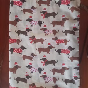 May include: Cream-colored fabric with a repeating pattern of dachshunds wearing Santa hats, scarves, and sweaters. The dachshunds are in various poses, with some wearing red and white polka-dot sweaters, others with snowflake patterns, and some with red scarves.