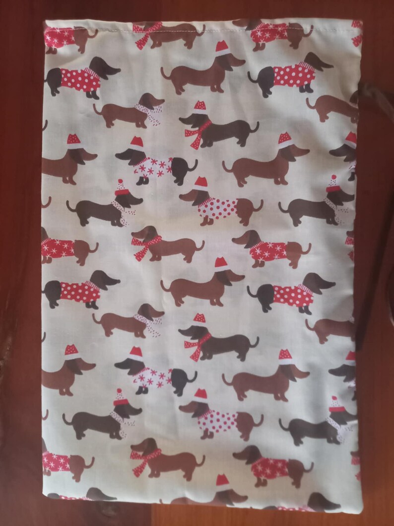 May include: Cream-colored fabric with a repeating pattern of dachshunds wearing Santa hats, scarves, and sweaters. The dachshunds are in various poses, with some wearing red and white polka-dot sweaters, others with snowflake patterns, and some with red scarves.