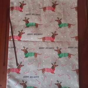 May include: A light gray fabric bag with a repeating pattern of dachshunds wearing reindeer antlers and sweaters in red and green. The words "HAPPY HOLIDAYS" are printed on the bag, along with candy cane designs.