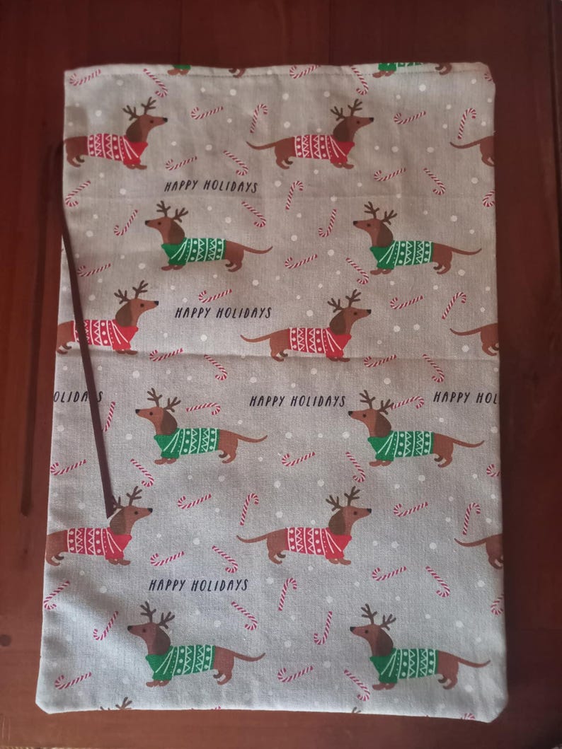 May include: A light gray fabric bag with a repeating pattern of dachshunds wearing reindeer antlers and sweaters in red and green. The words "HAPPY HOLIDAYS" are printed on the bag, along with candy cane designs.