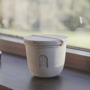 Peut inclure: Un pot à bougie en béton gris avec un couvercle et une corde attachée autour. Le pot a un motif découpé d'une plante dans un pot.