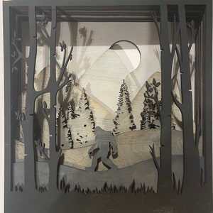 May include: A black silhouette of Bigfoot walking through a forest with a mountain in the background. The scene is framed by a black wooden shadow box.