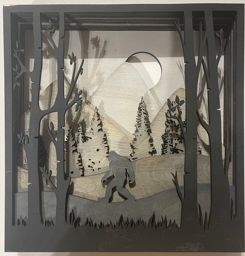May include: A black silhouette of Bigfoot walking through a forest with a mountain in the background. The scene is framed by a black wooden shadow box.