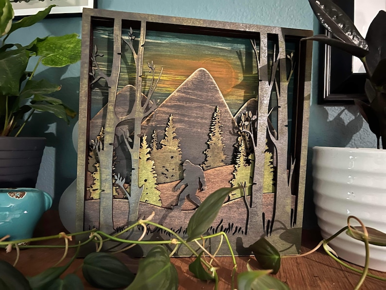 May include: A wooden wall art piece with a layered design depicting a forest scene with a silhouette of Bigfoot walking through the trees. The background is a sunset with a mountain range.