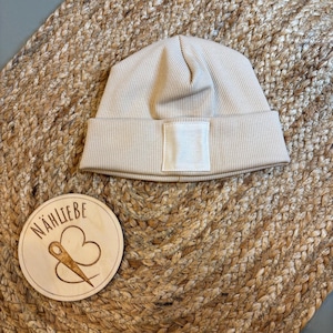 Könnte beinhalten: Eine beige gerippte Strickmütze mit einem weißen quadratischen Aufnäher auf der Vorderseite. Die Mütze wird auf einer gewebten, beigefarbenen Oberfläche präsentiert. Eine Holzscheibe mit dem Text "NÄHLIEBE" und einem Nadel- und Faden-Design ist ebenfalls sichtbar.