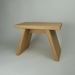 Könnte beinhalten: Ein einfacher Holzhocker mit rechteckiger Oberseite und einem einzigartigen A-Rahmen-Design. Der Hocker ist aus hellbraunem Holz gefertigt und steht auf einem weißen Hintergrund.
