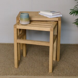 Könnte beinhalten: Ein Satz von zwei Holz-Nesting-Tischen mit hellem Eichenfinish. Eine Keramikvase mit Blau-, Grün- und Rosatönen steht auf dem oberen Tisch, zusammen mit einem Stapel Bücher. Die Tische sind rechteckig mit Lattenrosten. Höhe ca. 40 cm.