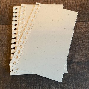 May include: A stack of off-white, textured paper sheets with a spiral binding on the left edge. The right edge of the paper has a decorative, uneven cut. The paper has small flecks throughout, giving it a natural appearance.