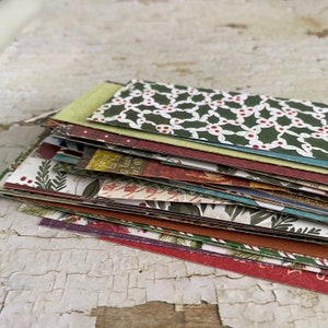 May include: A pile of patterned paper with festive Christmas designs. The papers feature holly, floral motifs, and text in green, red, and brown hues. The layered arrangement creates a textured appearance, suitable for crafting or scrapbooking.