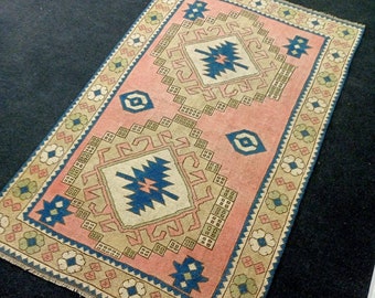 May include: A pink and beige rug with a traditional Turkish design. The rug features a central diamond shape with intricate geometric patterns in shades of blue, beige, and white. The border of the rug is decorated with floral motifs.