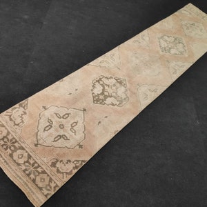 Könnte beinhalten: Ein langer, rechteckiger Teppich mit beigefarbenem Hintergrund und einem Muster aus braunen Rauten und floralen Motiven. Der Teppich hat einen Fransenrand.