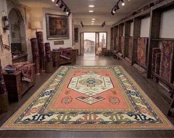 Puede incluir: Una alfombra rectangular grande con un campo de color salmón y un medallón central de color crema. La alfombra presenta patrones geométricos en azul marino, verde oliva y crema, enmarcados por un borde multicapa con complejos motivos lineales.