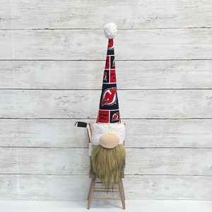 May include: A decorative gnome figurine with a long, pointed hat featuring the New Jersey Devils logo. The gnome has a beige nose, a long brown beard, and a hockey stick. It is sitting on a small wooden chair.