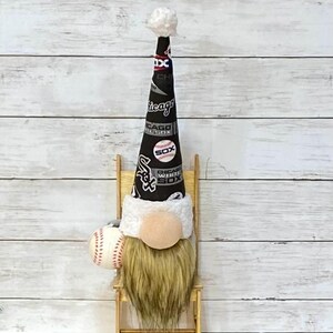 Könnte beinhalten: Ein Zwerg mit einem schwarz-weißen Baseball-Hut mit der Aufschrift "Chicago White Sox" und einem Baseball in der Hand. Der Zwerg sitzt auf einem kleinen Holzstuhl vor einem weißen Holz Hintergrund.