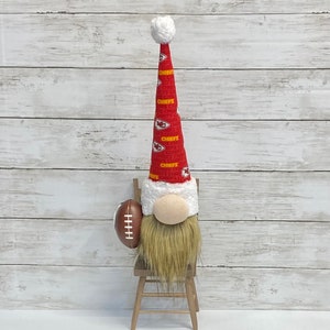 May include: A gnome wearing a red and yellow Kansas City Chiefs hat and holding a brown football. The gnome is sitting on a small wooden chair against a white wood background.