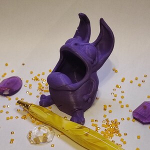 Könnte beinhalten: Eine violette 3D-gedruckte Figur eines Comic-Charakters mit weit offenem Mund. Die Figur sitzt auf einer weißen Oberfläche mit goldenem Glitzer und einem gelben Stift.