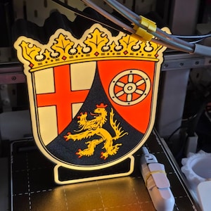Könnte beinhalten: Ein farbenfrohes, beleuchtetes Emblem mit Wappen-Design. Es zeigt eine goldene Krone, ein rotes Kreuz und einen goldenen Löwen auf schwarzem Hintergrund. Das Emblem befindet sich auf einem schwarzen Sockel mit weißem Rand und wird von hinten beleuchtet.