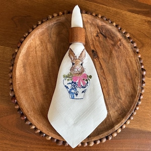 May include: A white linen napkin featuring an embroidered bunny design, held by a wooden napkin ring. The bunny is in a blue and white vase with a pink bow. The napkin is on a wooden plate with a beaded edge.