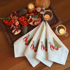 Puede incluir: Cuatro servilletas de lino blanco con diseños bordados de conejos y árboles de Navidad. Las servilletas están dobladas y dispuestas en una bandeja de madera con granadas, bayas, piñas y velas encendidas.