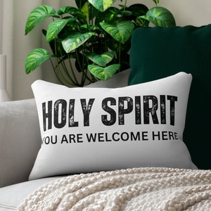 May include: White throw pillow with black text that reads "HOLY SPIRIT YOU ARE WELCOME HERE". The pillow is on a white couch with a green pillow and a white blanket.