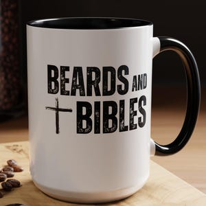 Puede incluir: Una taza de cerámica blanca con interior y asa negros. La taza presenta el texto "BEARDS AND BIBLES" en negrita negra con un símbolo de cruz. La taza está sobre una superficie de madera con una cuchara y un macarrón.