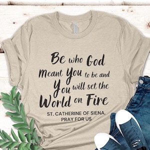 Puede incluir: Camiseta beige con el texto "Be who God Meant You to be and You will set the World on Fire" y "ST. CATHERINE OF SIENA, PRAY FOR US".