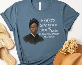 Op de afbeelding: Een blauw t-shirt met een portret van St. Josephine Bakhita en de tekst "IN GOD'S Will THERE IS GREAT Peace ST. JOSEPHINE BAKHITA PRAY FOR US." Het shirt is gestyled met geweven sandalen en een bruine hoed.