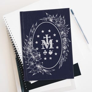 May include: A navy blue journal with a white floral design and an oval emblem featuring a cross and the letter "M" surrounded by stars. The journal is on top of a stack of notebooks and a pen.