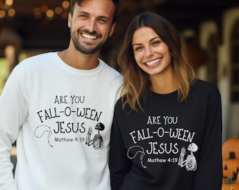 May include: A white long-sleeve sweatshirt and a black long-sleeve sweatshirt. Both shirts have the text "ARE YOU FALL-O-WEEN JESUS Matthew 4:19" with a skeleton and rosary design. Pumpkins are in the background.