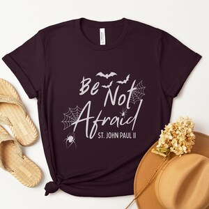 May include: A maroon t-shirt with the words "Be Not Afraid" and "St. John Paul II" in white script, along with bat and spider web graphics. The shirt is paired with woven sandals and a tan hat with a flower.