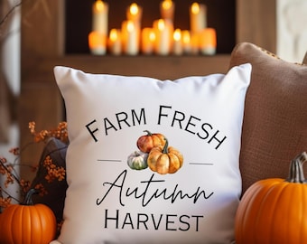 May include: A white pillow with the text "FARM FRESH AUTUMN HARVEST" and watercolor pumpkins. Two orange pumpkins and a small green pumpkin are next to the pillow. The background includes a fireplace and candles.