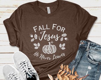 Puede incluir: Una camiseta marrón con el texto blanco "FALL FOR Jesus He Never Leaves", un diseño de calabaza y hojas. La camiseta se combina con vaqueros azules, zapatillas blancas y elementos de follaje otoñal.
