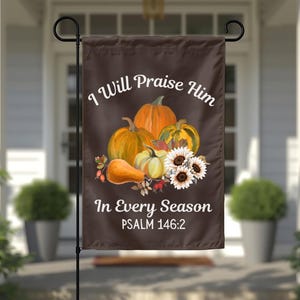 Puede incluir: Una bandera de jardín rectangular de color marrón con una ilustración central de calabazas naranjas, una calabaza tipo cacahuete y girasoles blancos. El texto "I Will Praise Him In Every Season" y "PSALM 146:2" aparece en letras blancas.