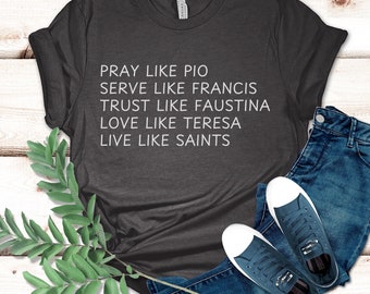 May include: A dark grey t-shirt with white text that reads "Pray like Pio, Serve like Francis, Trust like Faustina, Love like Teresa, Live like Saints". The shirt is laid out on a light wood surface with a sprig of green leaves and a pair of blue jeans and blue sneakers.